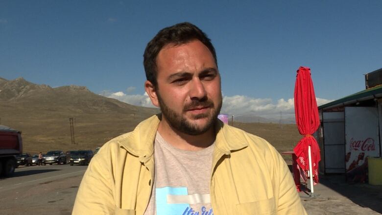 A man in a yellow shirt stands in front of a busy road in a mountainous region.