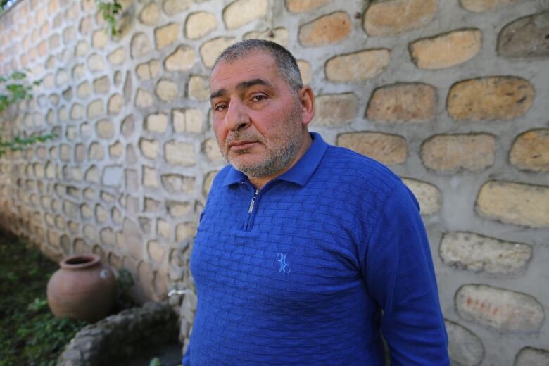 An older man in a blue shirt stands solemnly in front of a wall.