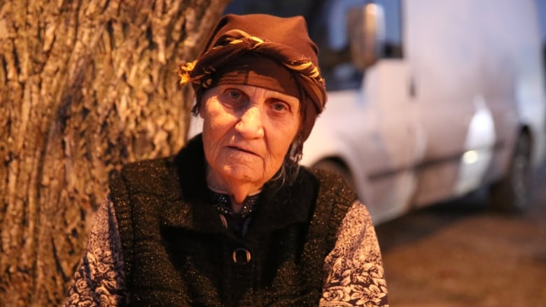 An old woman in traditional dress sits by a tree.