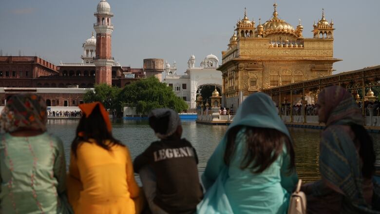 People sit along a wall beside a body of water with a golden temple in the background.