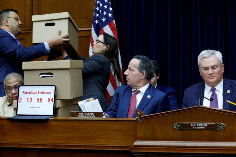 Three boxes behind members of Congress, seated. In foreground, a clock saying a government shutdown starts in two days.