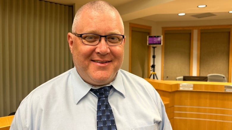 A man in a light coloured dress shirt with a blue checked tie wearing glasses and smiling.