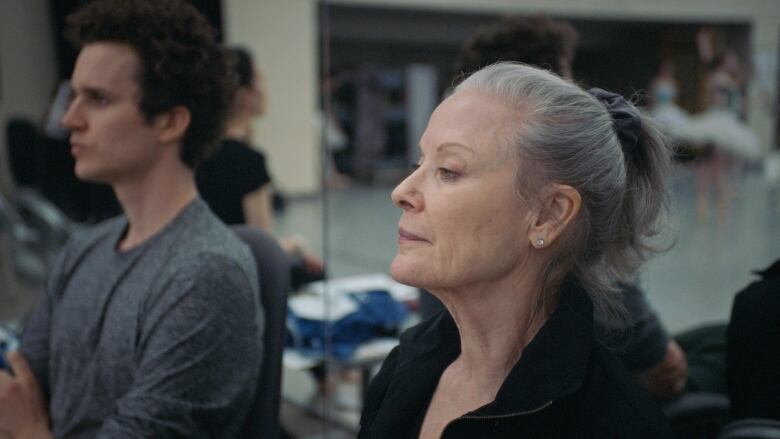 A woman sitting next to a man -- both in front of a mirrored wall -- looks out of frame. 