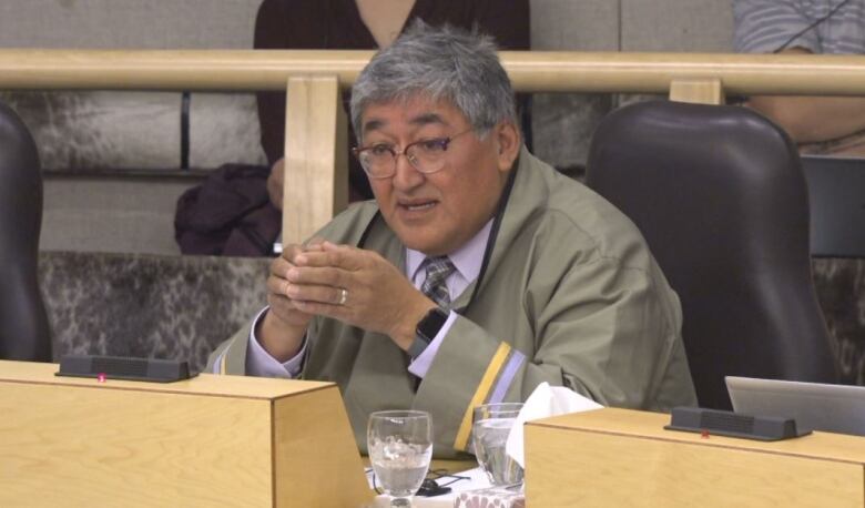 A man with grey-ish hair sits in the Nunavut legislative assembly and speaks to members. 