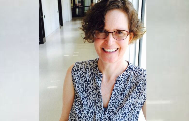 A woman with short, dark curly hair and burgundy-rimmed glasses is pictured in a sleeveless, black and white textured blouse, smiling at the camera. 