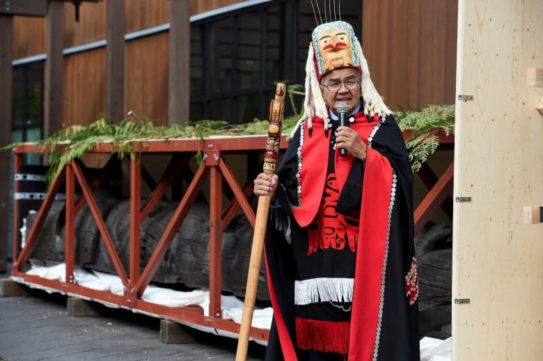 A man stands speaking in front of a large totel pole lying on its side. 