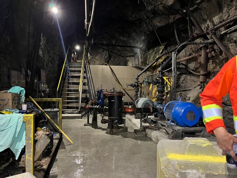 A rocky cavern with machines and pipes lining on side of the image and a shelf on the other. There's also a staircase leading up over a wall.