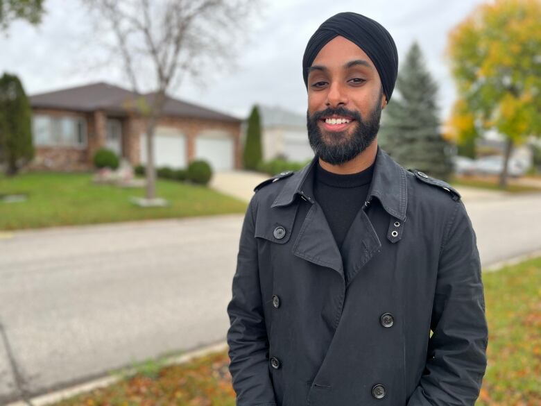 A man wearing a head covering and a black coat stands next to a residential street.