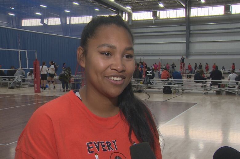 Veronica Headley smiles wearing an orange shirt while she stands in front of a volleyball court.