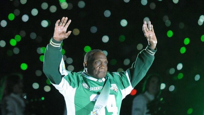 A man wearing a football jersey waves to fans in the stands with both arms in the air.