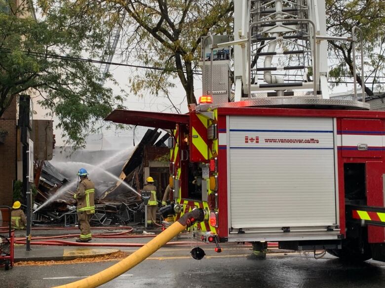 Three firefighters spray water from firehoses on a damaged building, with a firetruck in the foreground. 