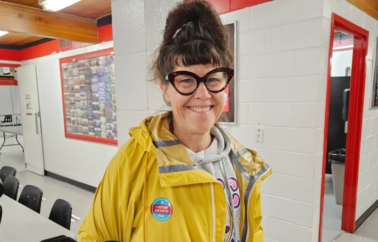 A woman with her hair in a tall bun, and wearing large bold glasses and a yellow raincoat, smiles broadly at the camera.