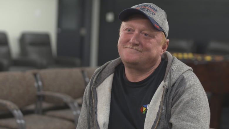 A strawberry blonde-haired man is smiling and is wearing a hoodie and baseball cap. 