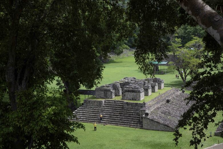 Ancient ruins are seen from a distance through tree branches. A handful of people are seen scattered throughout the site.