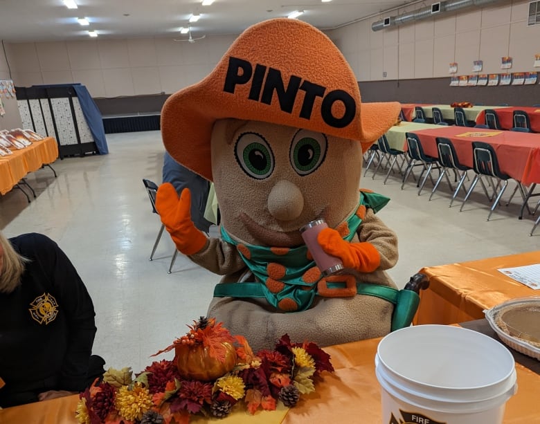 A person in a costume waves at the camera. It's a bean in a orange hat which reads 'Pinto.'