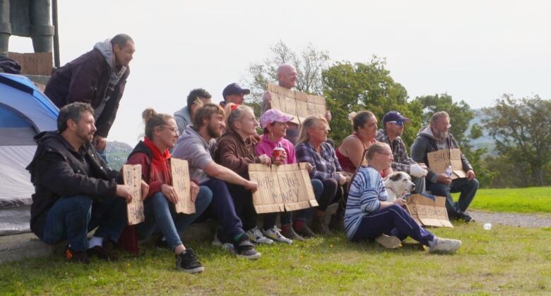 A group of people sit on a grassy field holding cardboard signs.