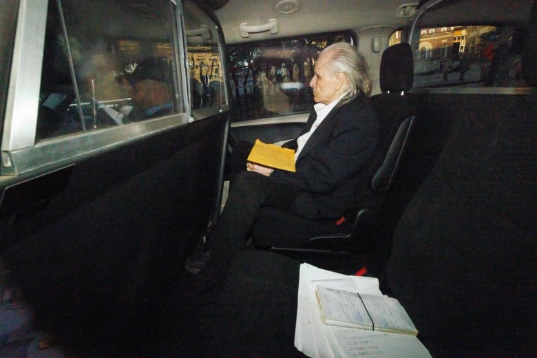 An older man with white hair, wearing a black suit and white shirt, sits with his legs crossed in the back of a vehicle.