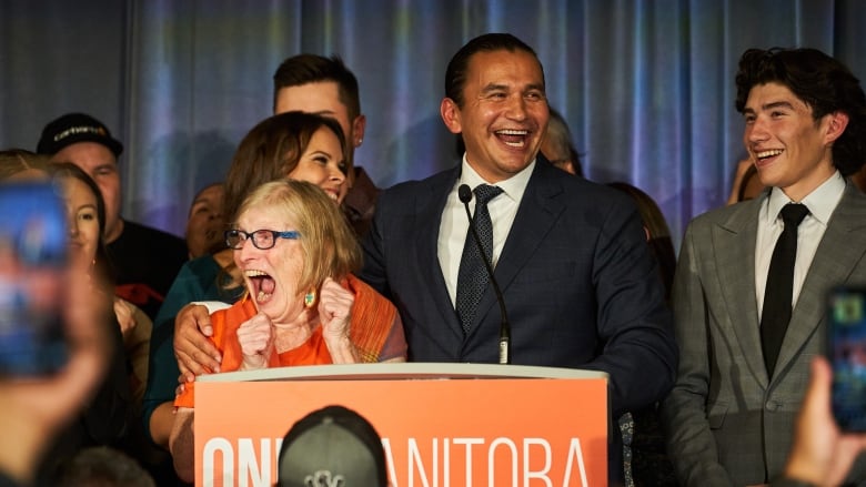 A smiling man in ablue suit puts his arm around a woman in an orange dress as they both stand behind a podium.