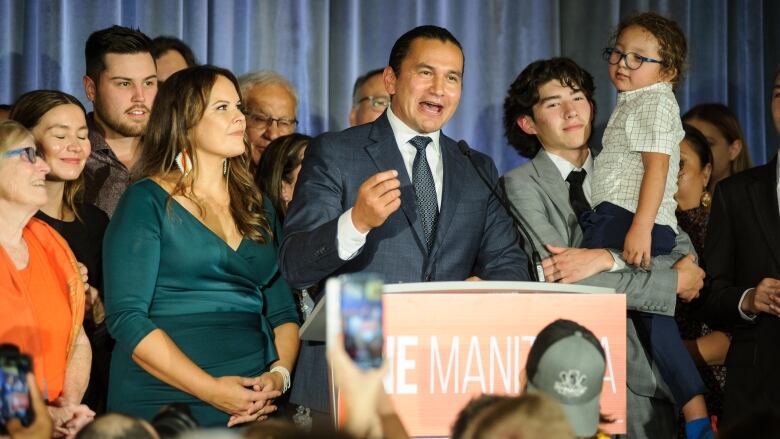 A man in a blue suit surrounded by people speaks at a podium.