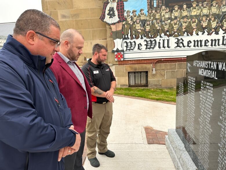 Three men are shown looking at a black stone monument which has the names of 158 Canadian soldiers who died in Afghanistan etched in white writing. 