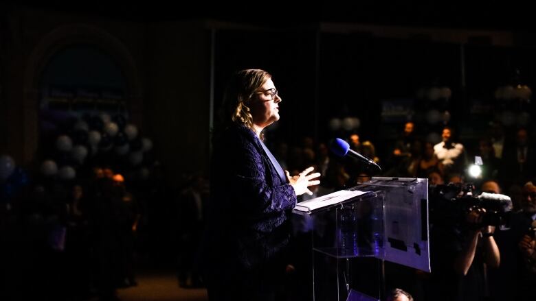 A woman stands behind a podium, speaking. She's lit up against a dark room.