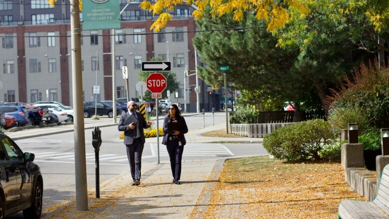 Two people in suits walk down a sidewalk on a leafy thru street.