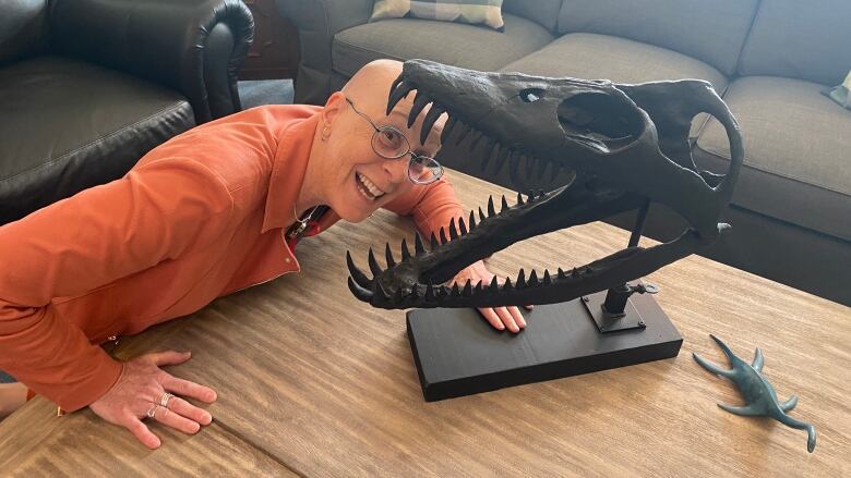 A bald woman poses her head to appear as if a fossil cast is eating her on a table.