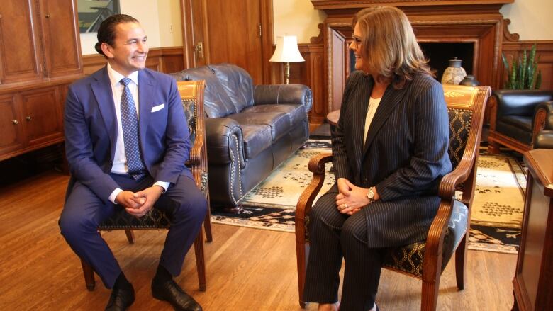 A man in a blue suit and tie is seated beside a woman in a blazer with white stripes.