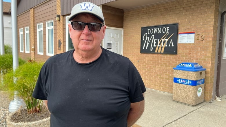 A man wearing a black t-shirt, black sunglasses and a grey hat with the letter 'W' on it stands in front of a brick building with a sign on it saying Town of Melita.