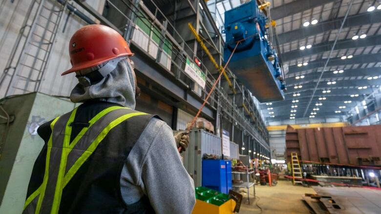 A man is seen guiding a large engine overhead in a large ship construction hangar.