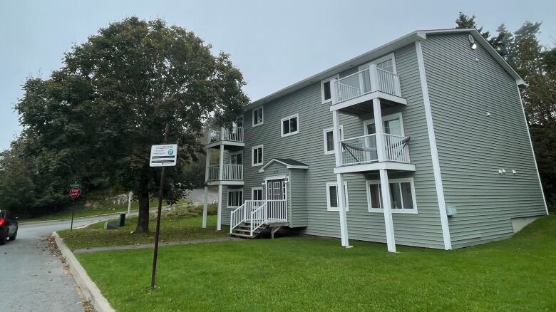 A small grey apartment building with white trim sits on a grassy corner.