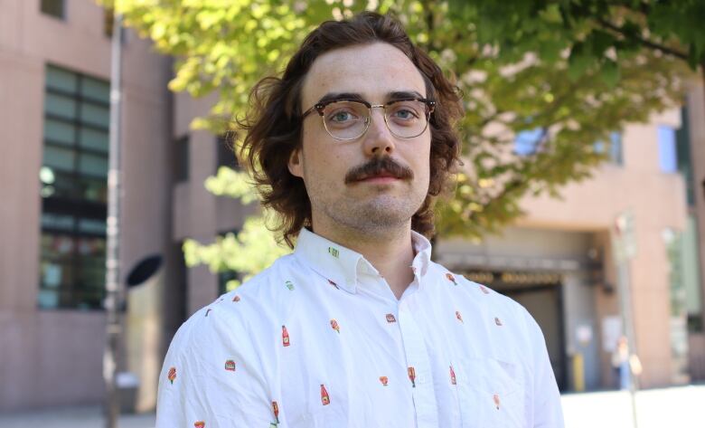 A long haired and mustached white man is looking at the camera, wearing a white shirt. 