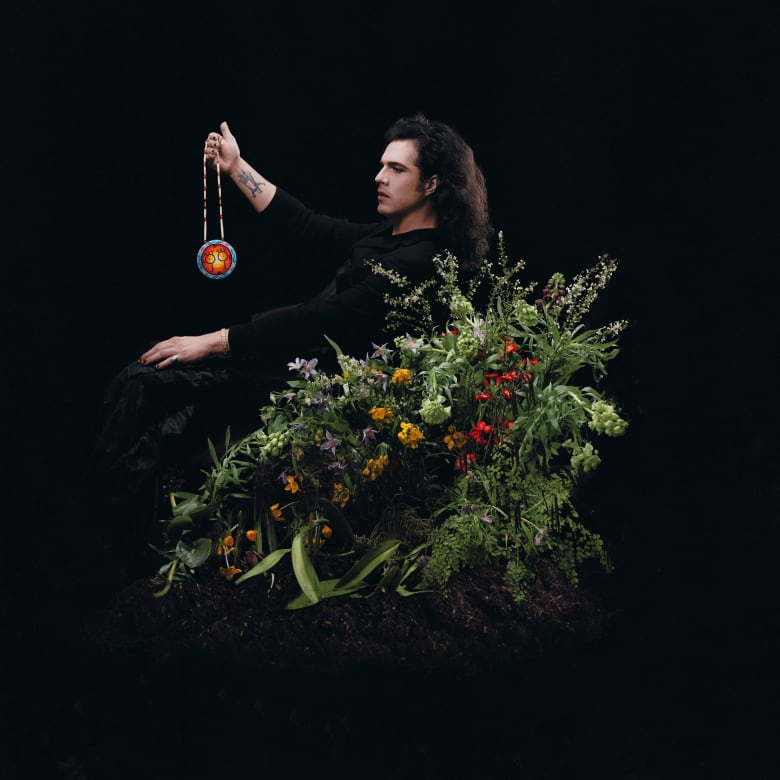 Side profile of musician Jeremy Dutcher holding a beaded necklace, surrounded by flowers. 