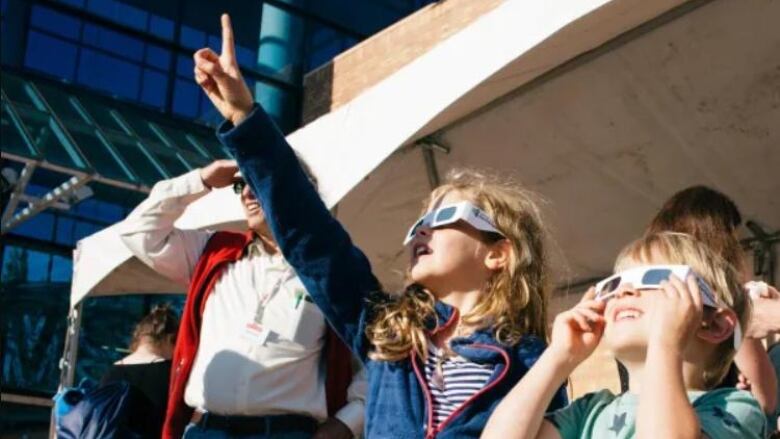 Two kids, a girl about 9 years old on the left, with a boy about 7 years old on the right, wear protective eye gear as they look up and point towards the sky.