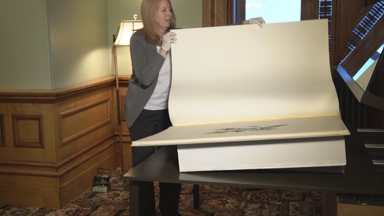 A woman in a grey sweater turns the page on a massive book that sits in a case.