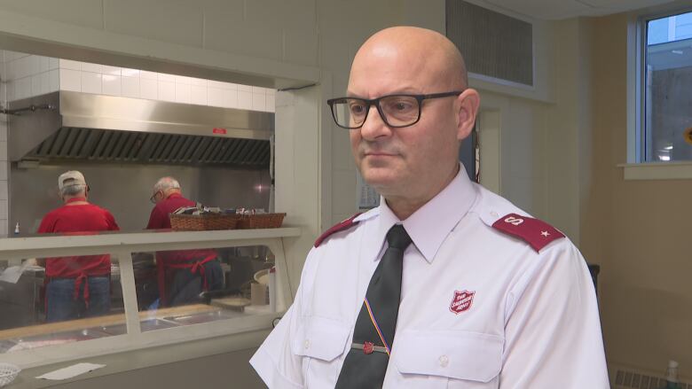 A man in a Salvation Army uniform stands. Behind him, people work on a kitchen.