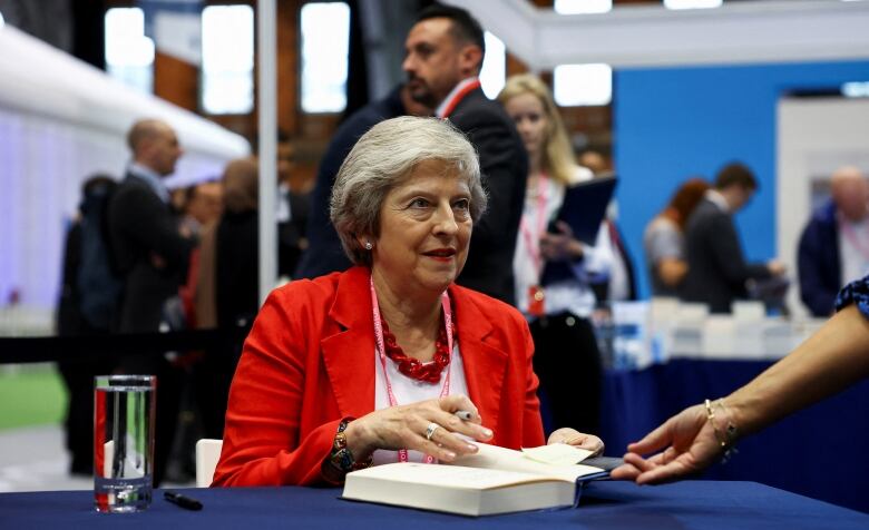 A woman with short grey hair, wearing a white top and a red jacket, signs a book.