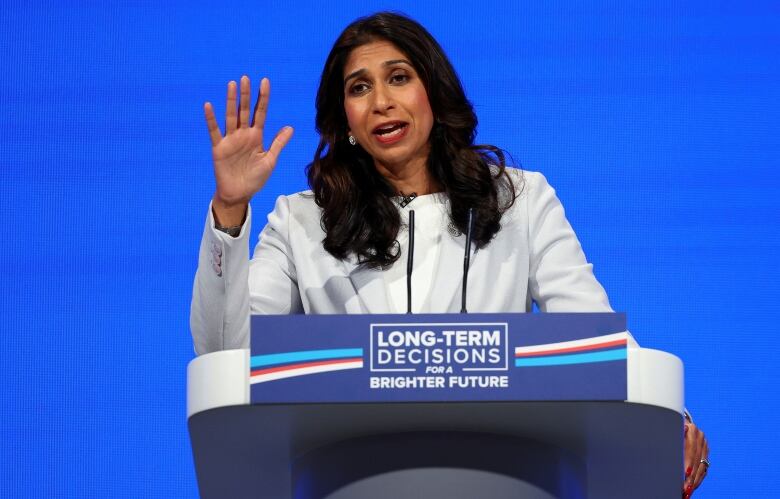 A woman with long, dark hair, wearing a white suit, gestures as she speaks at a podium.