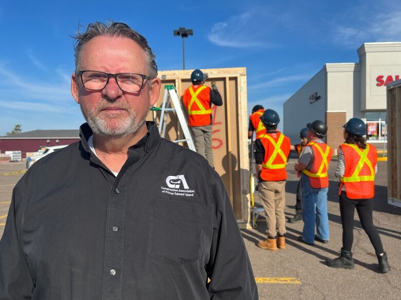 A man wearing glasses and a black button up shirt stands against a backdrop of apprentice workers practicing on a structure.