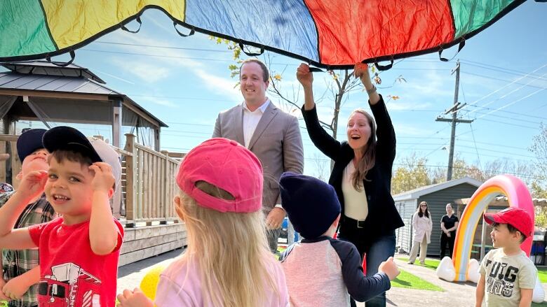 A woman smiles and lifts a multi-coloured parachute over the heads of excited young children as a man in a grey suit smiles and looks on.