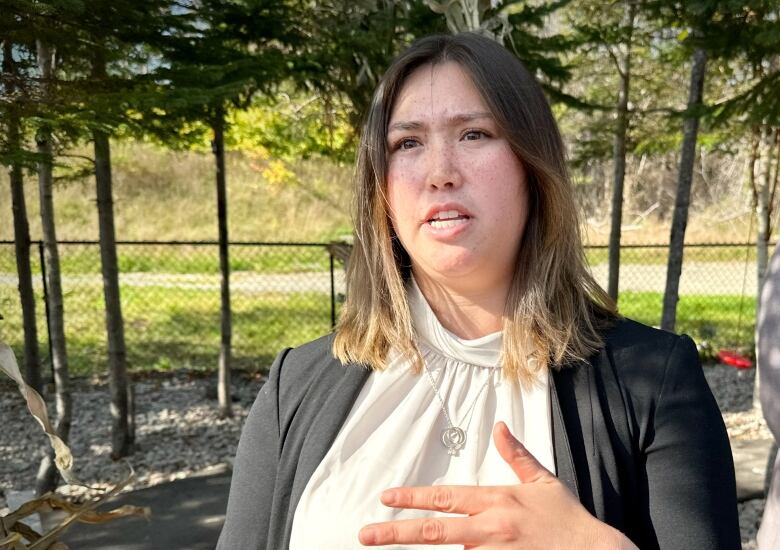 A woman with a grey cover over a white blouse speaks at an outdoor event with trees in the background.