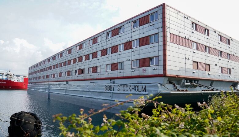 A long three-storey grey building with windows sits on a barge floating in the water.