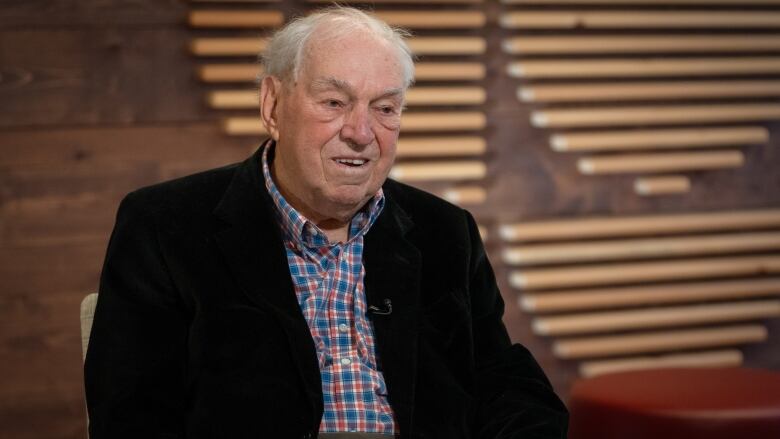 An old man in a plaid shirt and blazer sits in a studio, smiling. 