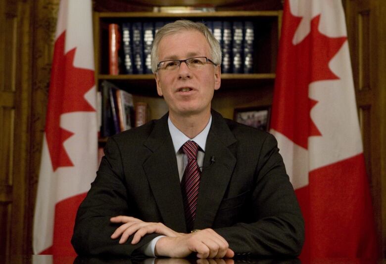 A man in a suit sits at a table, looking forward. Two Canadian flags are behind him. 