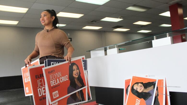 A woman stands in an empty room holding orange signs with her name on them.