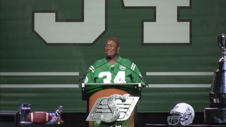 A black woman wearing a green football jersey, with the number 34 on the front, is standing at a podium, speaking into two microphones.