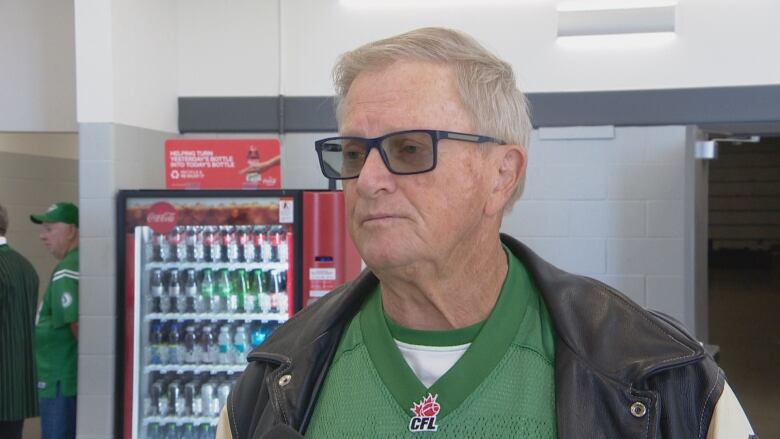 An older white man, with greying hair and glasses, is wearing a green football jersey underneath a leather jacket. Behind him is a Coca-Cola vending machine.