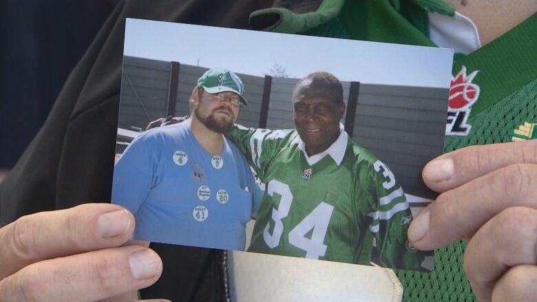 A white man with a brown beard, blue t-shirt and green ball cap is posing next to a Black man, who's wearing a green football jersey with the number 34 on the front. The Black man is on the right, and has his hand wrapped around the other man.