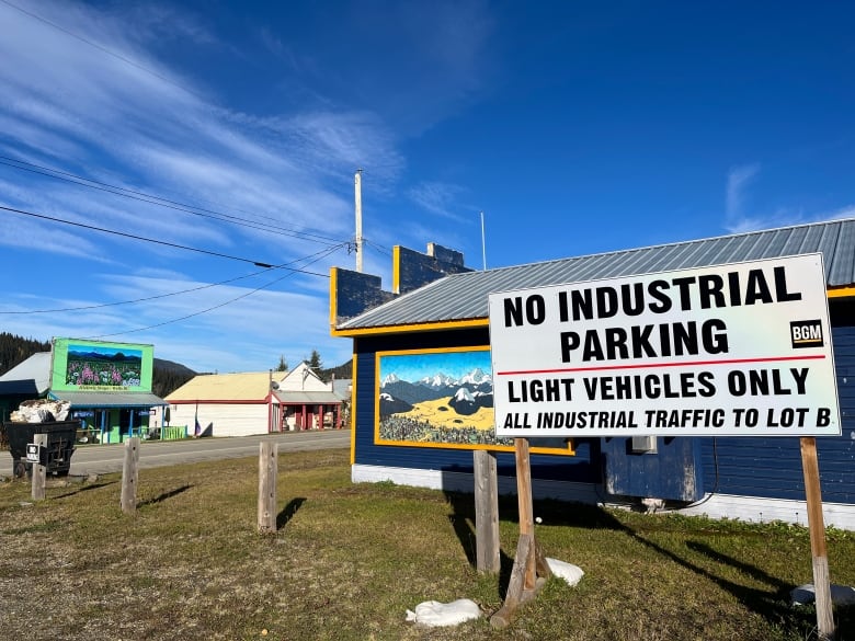 In a small town, historic buildings are seen with mountain murals painted on them, in front of which a sign says 'No Industrial Parking' and directs industrial traffic to a parking lot.