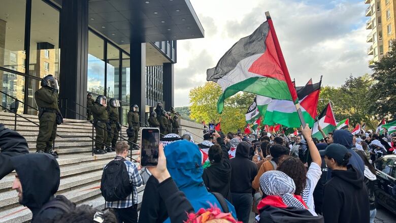 A crowd waving Palestinian flags gather at the Israeli consulate. Israeli guards stand on the steps of the building. 